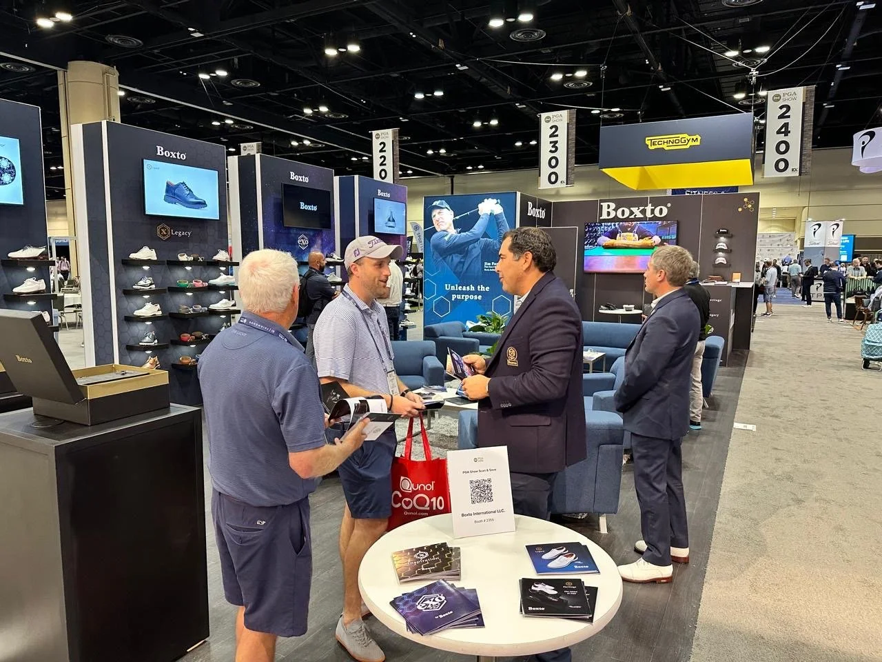 People talking at a booth exhibiting golf shoes and accessories at a trade show, with display shelves and banners behind them.