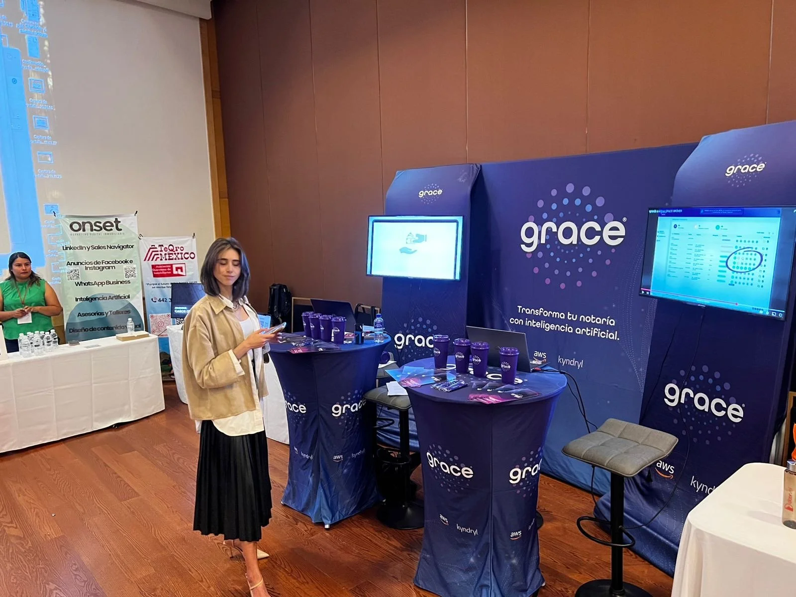 A woman standing in front of a blue booth at an event, with screens and promotional materials displayed, and another woman in the background near a table with bottled water.