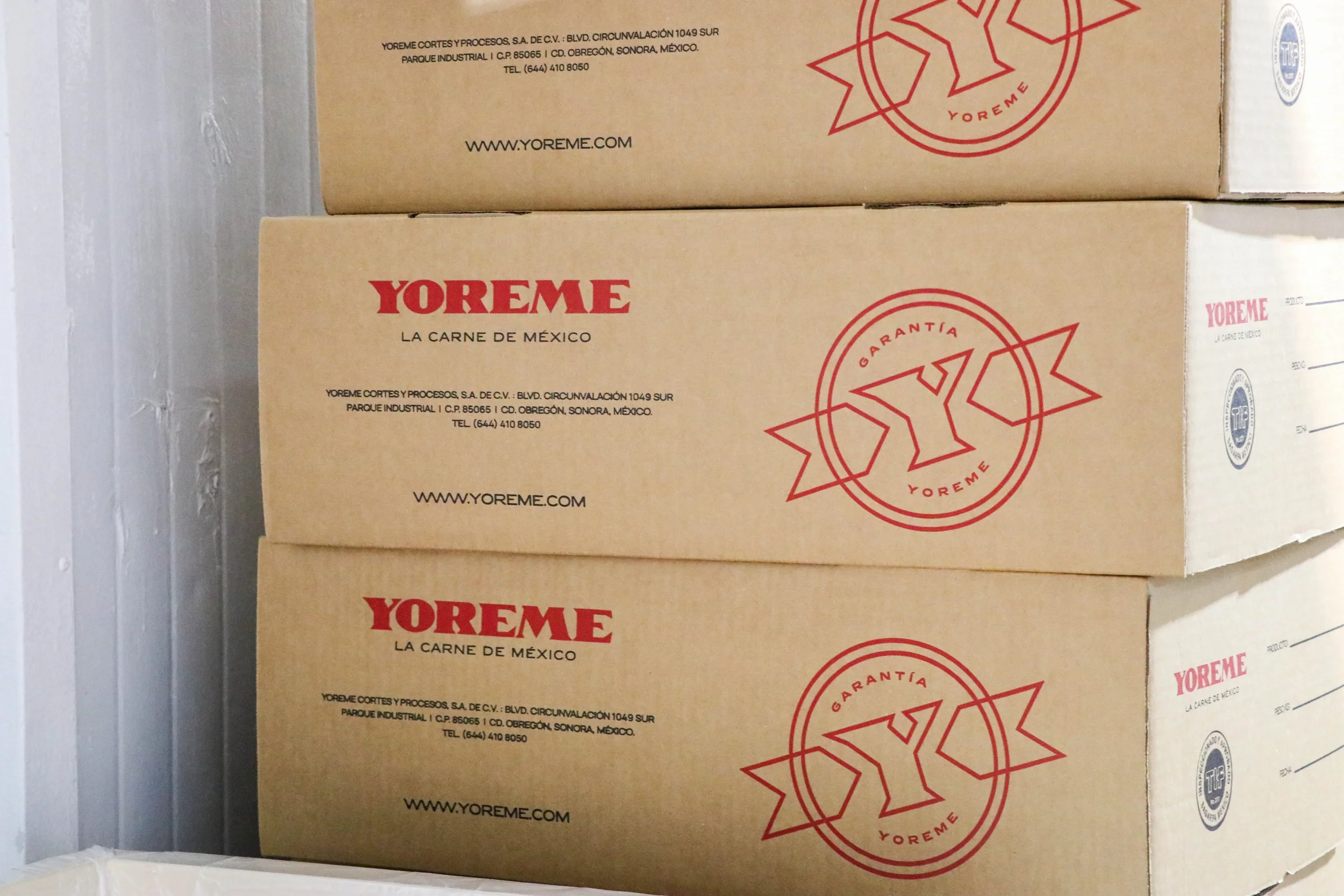 Stacked cardboard boxes labeled YOREME, a Mexican meat brand, with red Yoreme logos and website, stored against a wall.