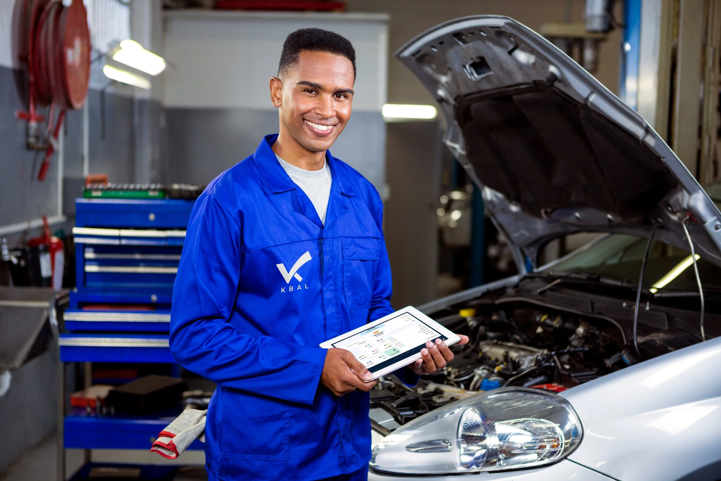 A smiling mechanic in a blue uniform holding a tablet standing next to an open car hood in an auto repair shop.
