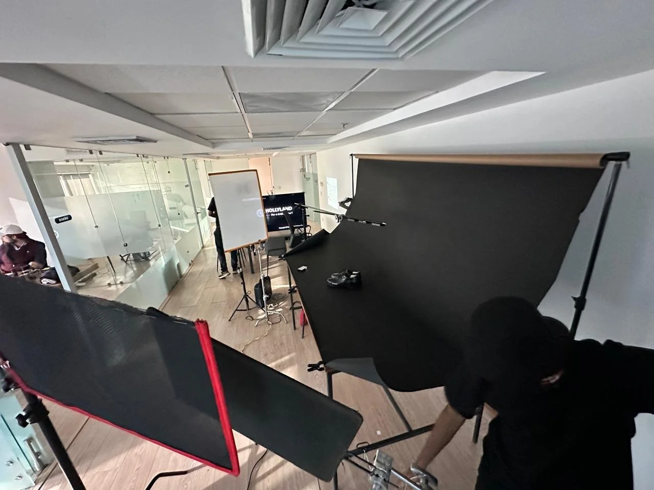 Inside a modern office with a photography setup including large black reflectors and a whiteboard, a person preparing for a photo shoot, and a background display that reads 'Holland.'