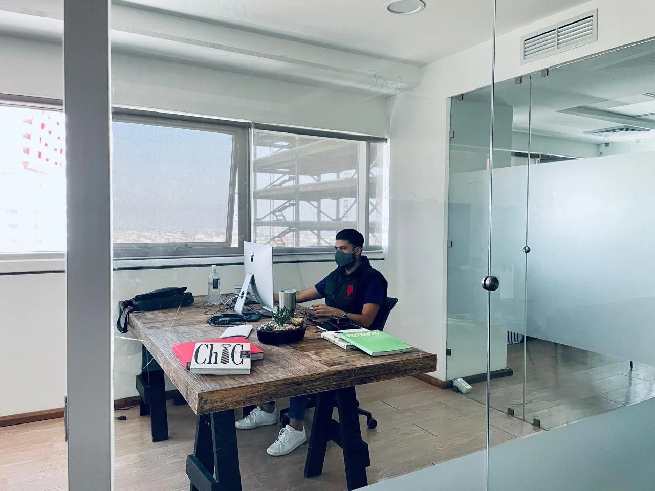 A man wearing a black face mask and a dark shirt sitting at a wooden desk in an office, working on a computer. The desk has a potted plant, a bottle of hand sanitizer, books, and a red box labeled "Chic." The office has large windows with a view of a