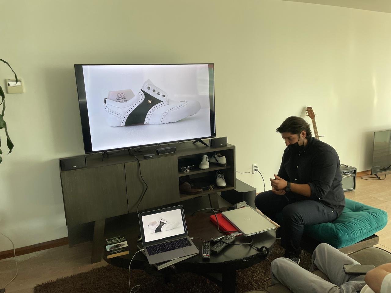 Person sitting on a green cushioned seat watching a presentation of a white and black sneaker on a large TV screen.