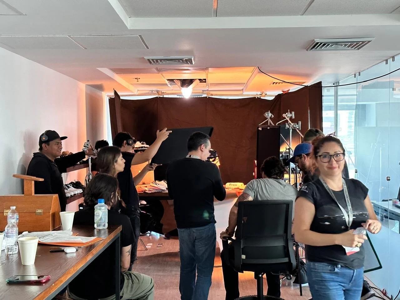 Behind-the-scenes of a photo shoot or filming, with crew members setting up equipment in a studio with a brown backdrop and lighting.