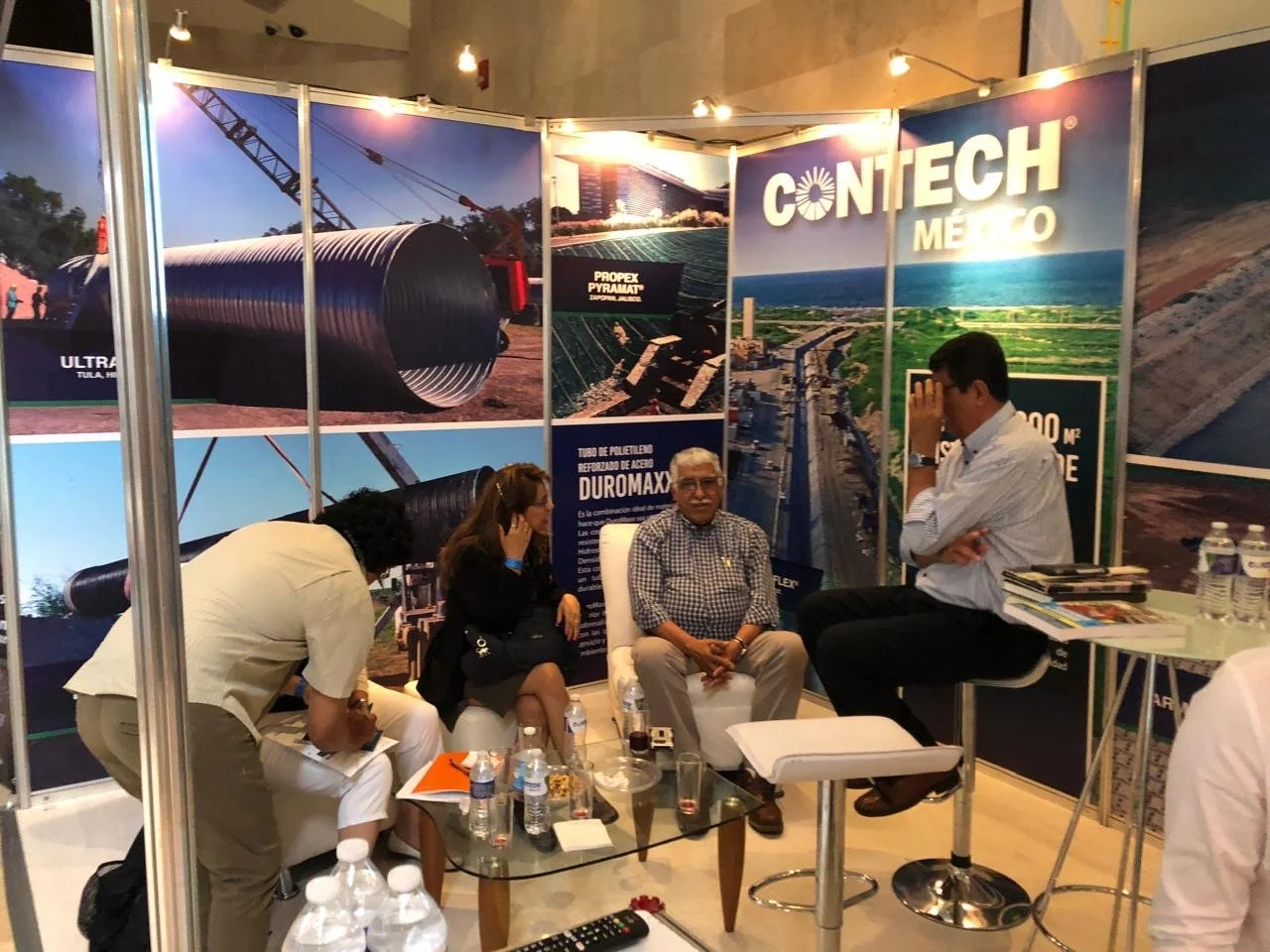 A trade show booth with three people having a conversation. The booth features large posters of construction equipment and infrastructure projects, with prominent text such as 'CONTECH MEXICO' and images of pipes, bridges, and construction sites.