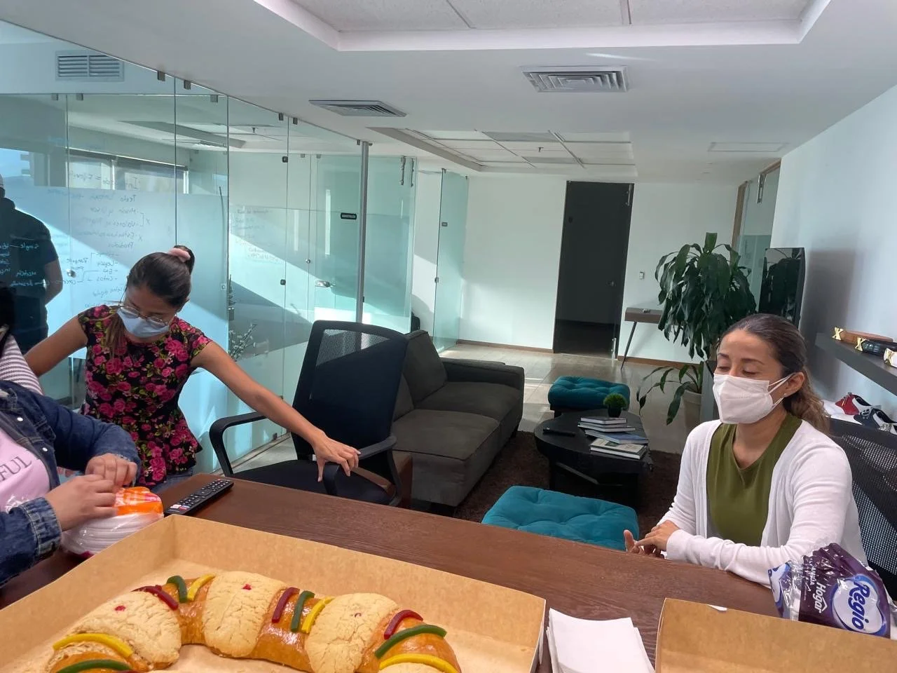 Office meeting room with three women wearing face masks. One woman is seated at the table, while another is reaching for a plastic bag. The room has a sofa, bookshelf with books, and a large plant. There are cookies with colorful candy on the table i
