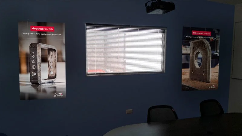 Office room with two posters advertising Kloeckner Metals, a window with blinds, and chairs around a table.