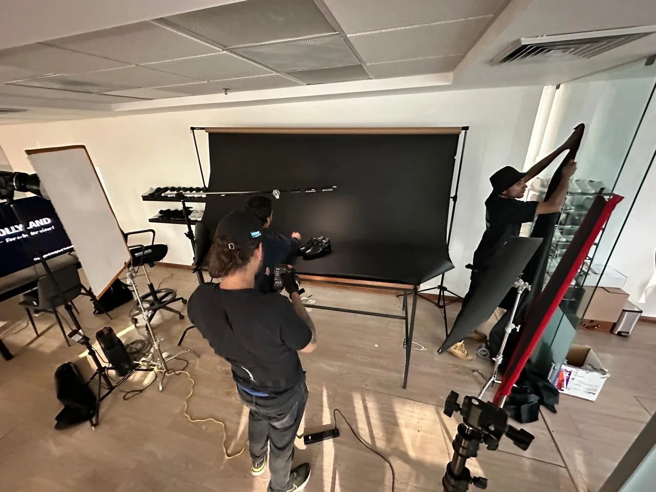 A photo of a video production setup with three crew members preparing for filming. There is a black backdrop, lighting equipment, and a camera on a tripod. One person is adjusting the backdrop, while another is holding a camera, and the third is arra