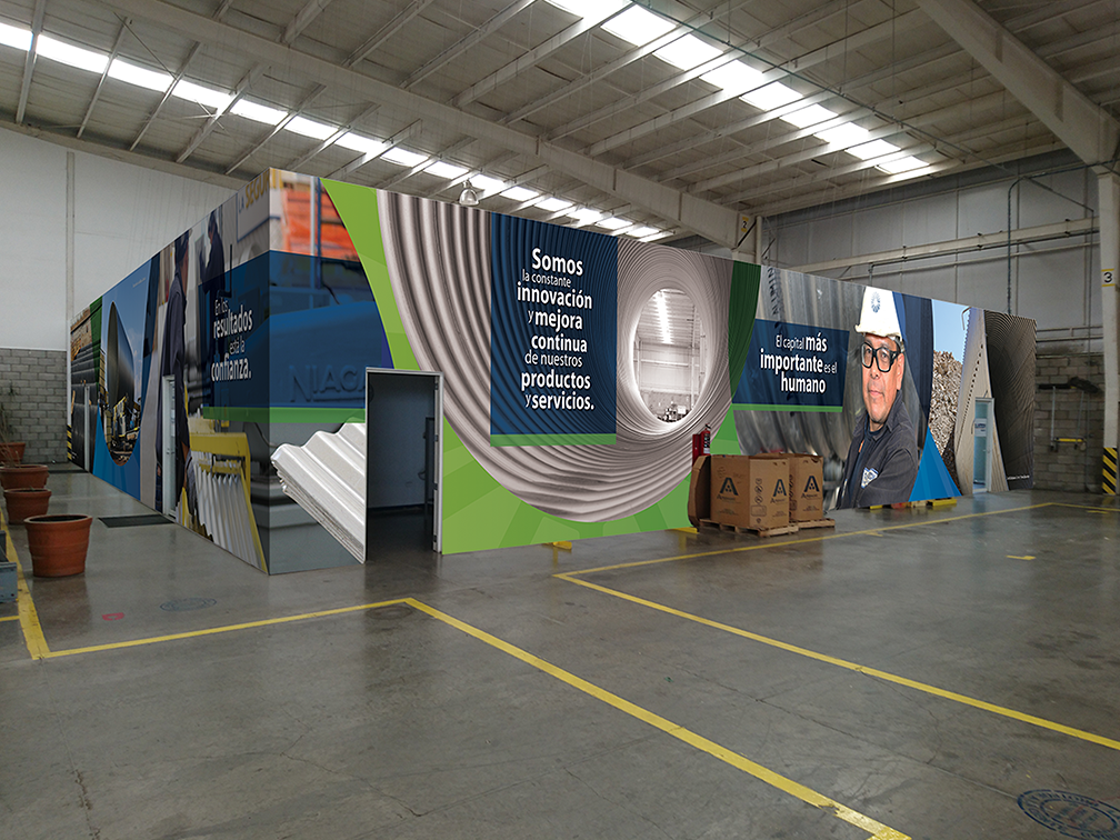 Large indoor space with a colorful, curved wall display featuring images and text about innovation and trust. The display includes graphics, a worker wearing safety gear, and bilingual text in Spanish and English. There are cardboard boxes and orange