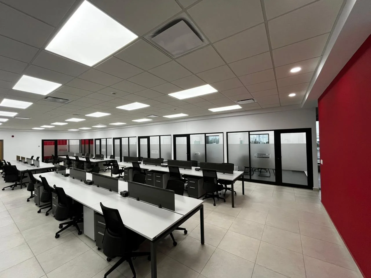 Empty modern office space with rows of black desks and chairs, glass-walled conference room, white ceiling with fluorescent and recessed lighting, beige tiled floor, one red accent wall, and large windows.