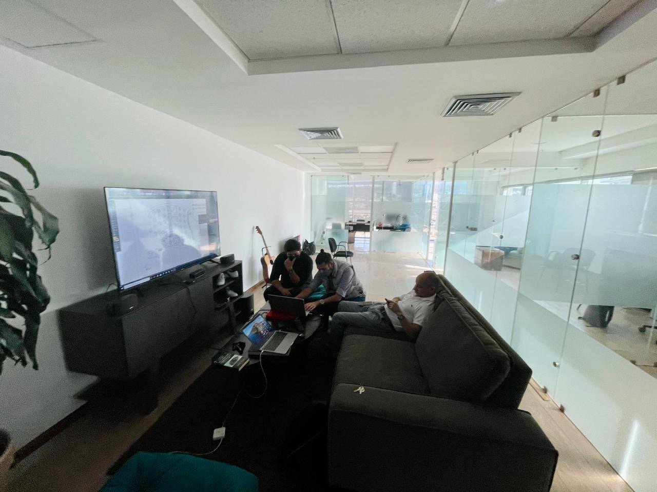Three people sitting in a modern office lounge area, two working on laptops and one on a smartphone, with a large flat-screen TV on the wall, glass walls with frosted designs, and office furniture in the background.