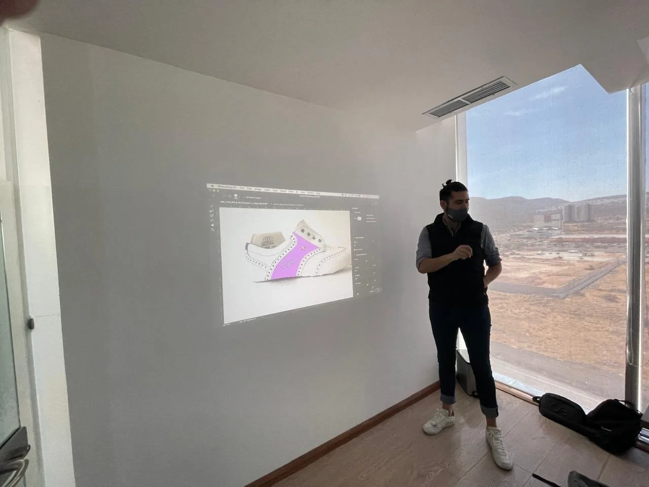 A person standing in a room with a projection of a design on the wall, showing a shoe with pink and purple details, during a presentation or discussion.