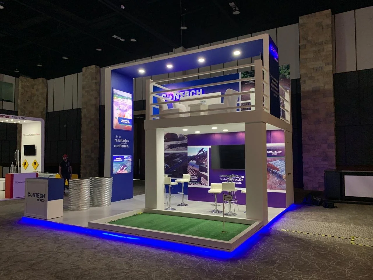 Exhibition booth with a two-story structure, featuring a lower level with white chairs and a high-top table, an upper balcony with white chairs, and blue lighting accents, at a construction industry trade show.