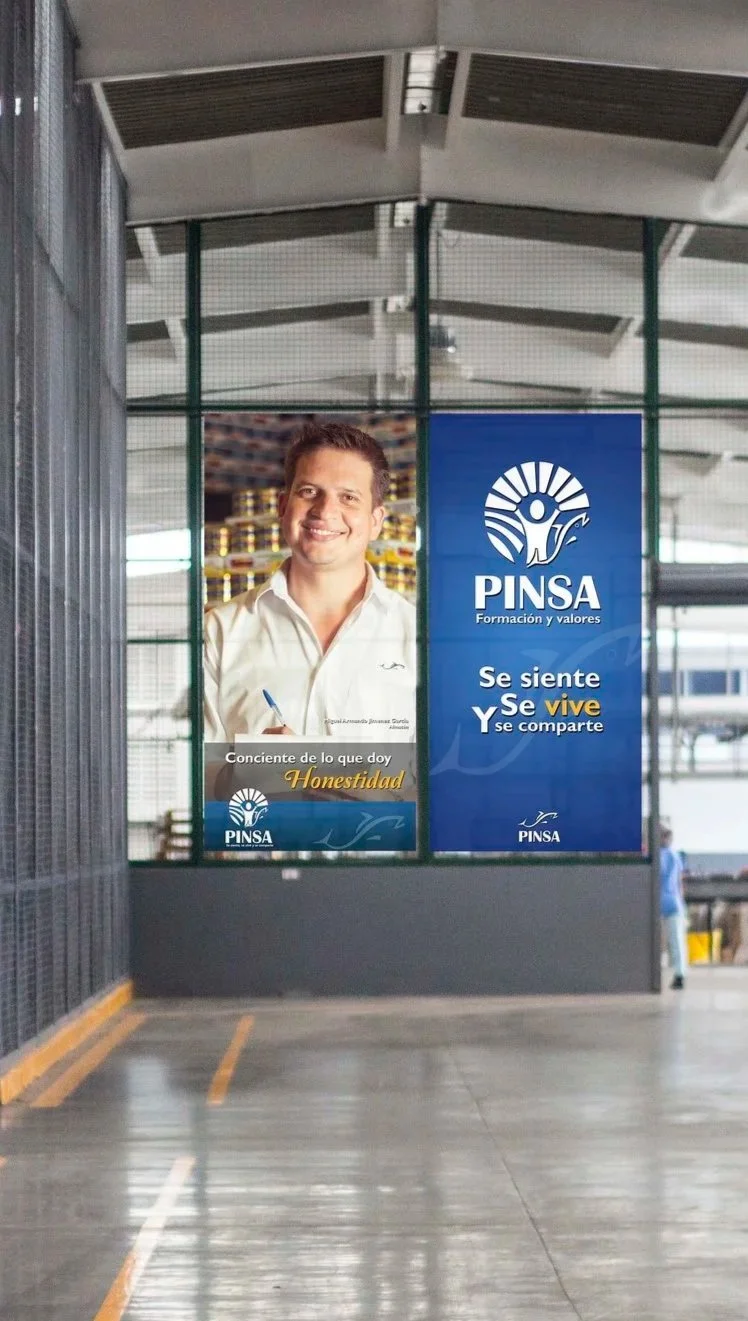 Large banner inside a building with a smiling man holding a pen, advertising for PINSA education institution. The banner includes the PINSA logo and slogan in Spanish, emphasizing honesty and sharing feelings.