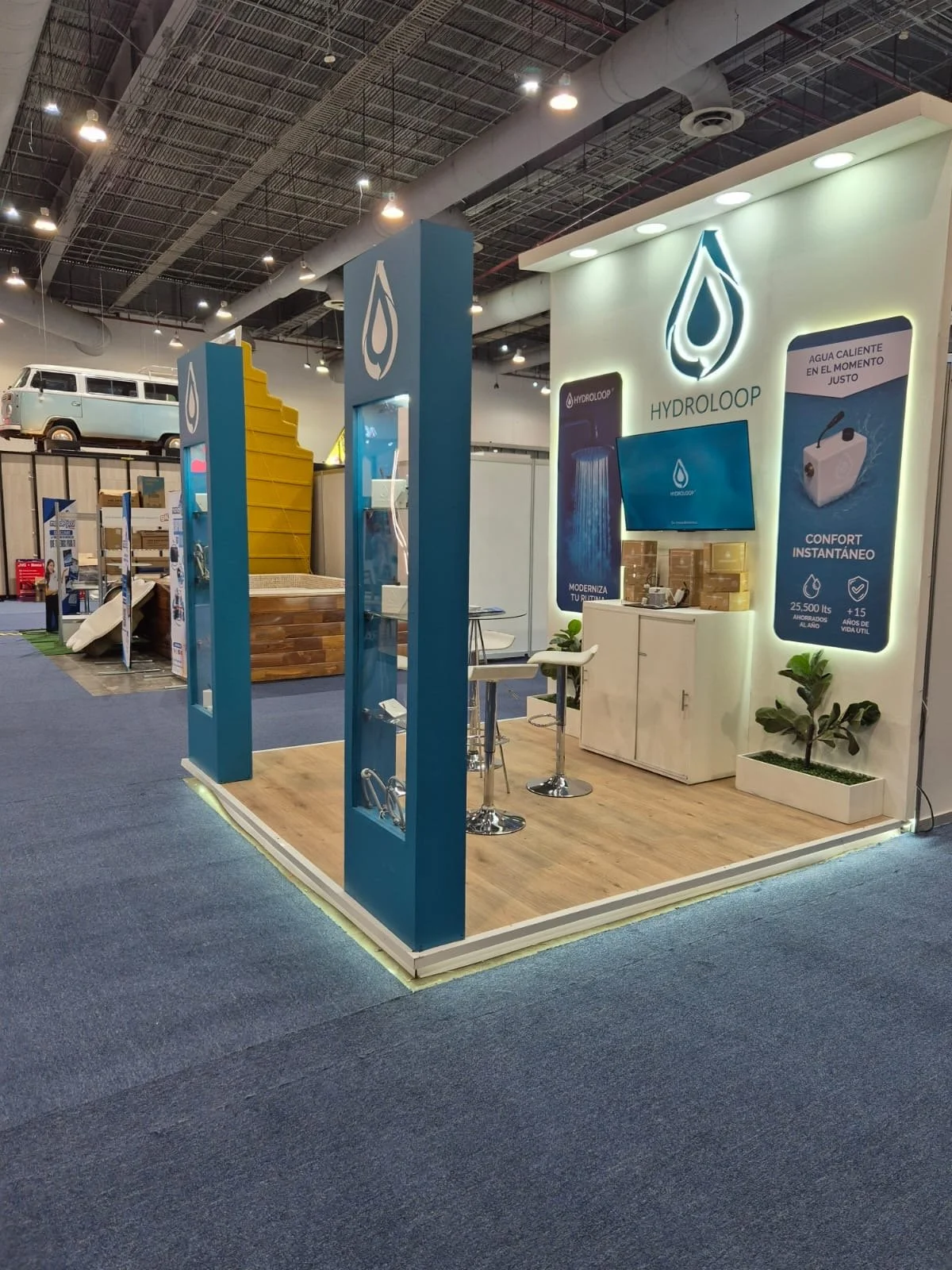 Exhibition booth for HydrolLoop featuring a branding logo, display screen, promotional posters, a small white cabinet, potted plants, and seating area with stools inside a convention center.