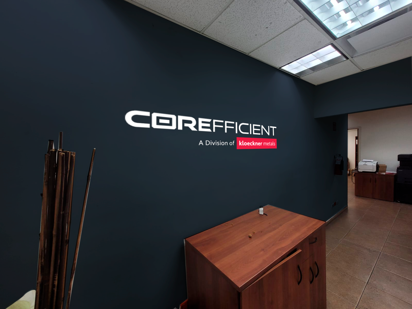 Office interior with a dark blue wall showing the logo of CORE efficient, a division of Kloeckner Metals. There is a wooden cabinet in front and some items in the background.