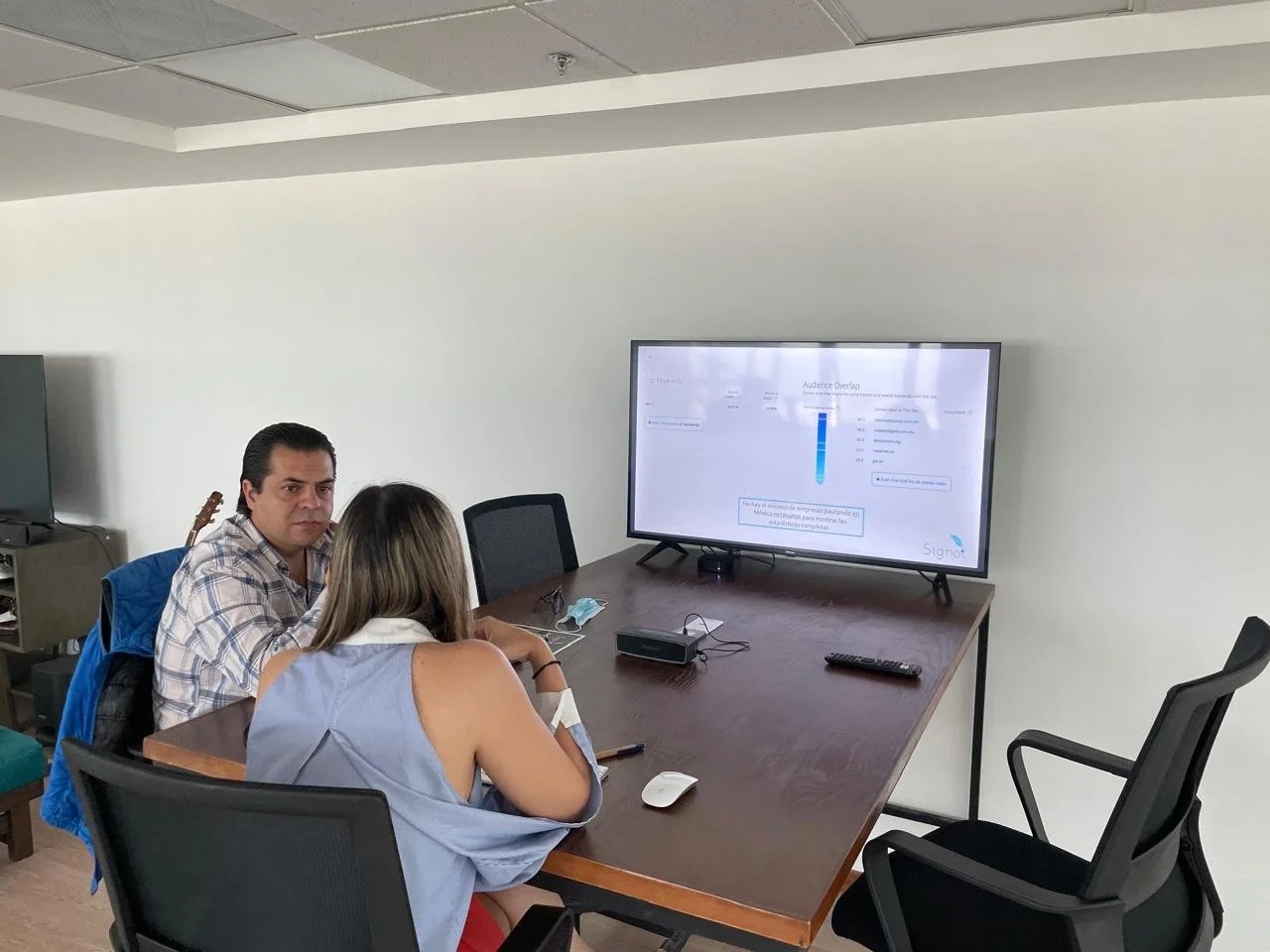 Two people sitting at a conference table looking at a large monitor displaying a presentation, with a remote and a mouse on the table.