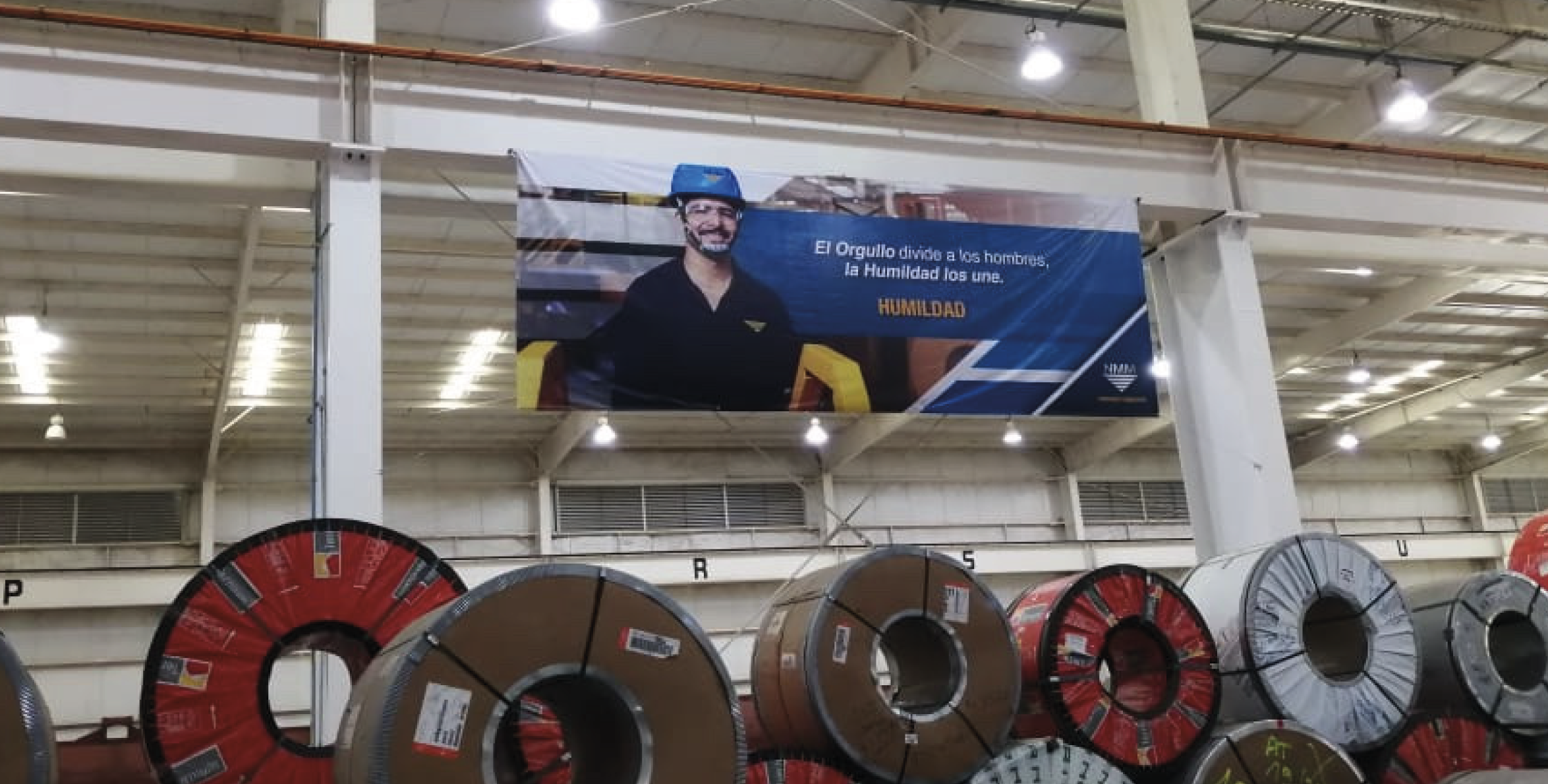 Industrial warehouse with large coils of metal on the floor and a poster hanging from the ceiling showing a smiling man wearing a blue hard hat and a black shirt. The poster includes the Spanish text: 'El Orgullo divide a los hombres; la Humildad los