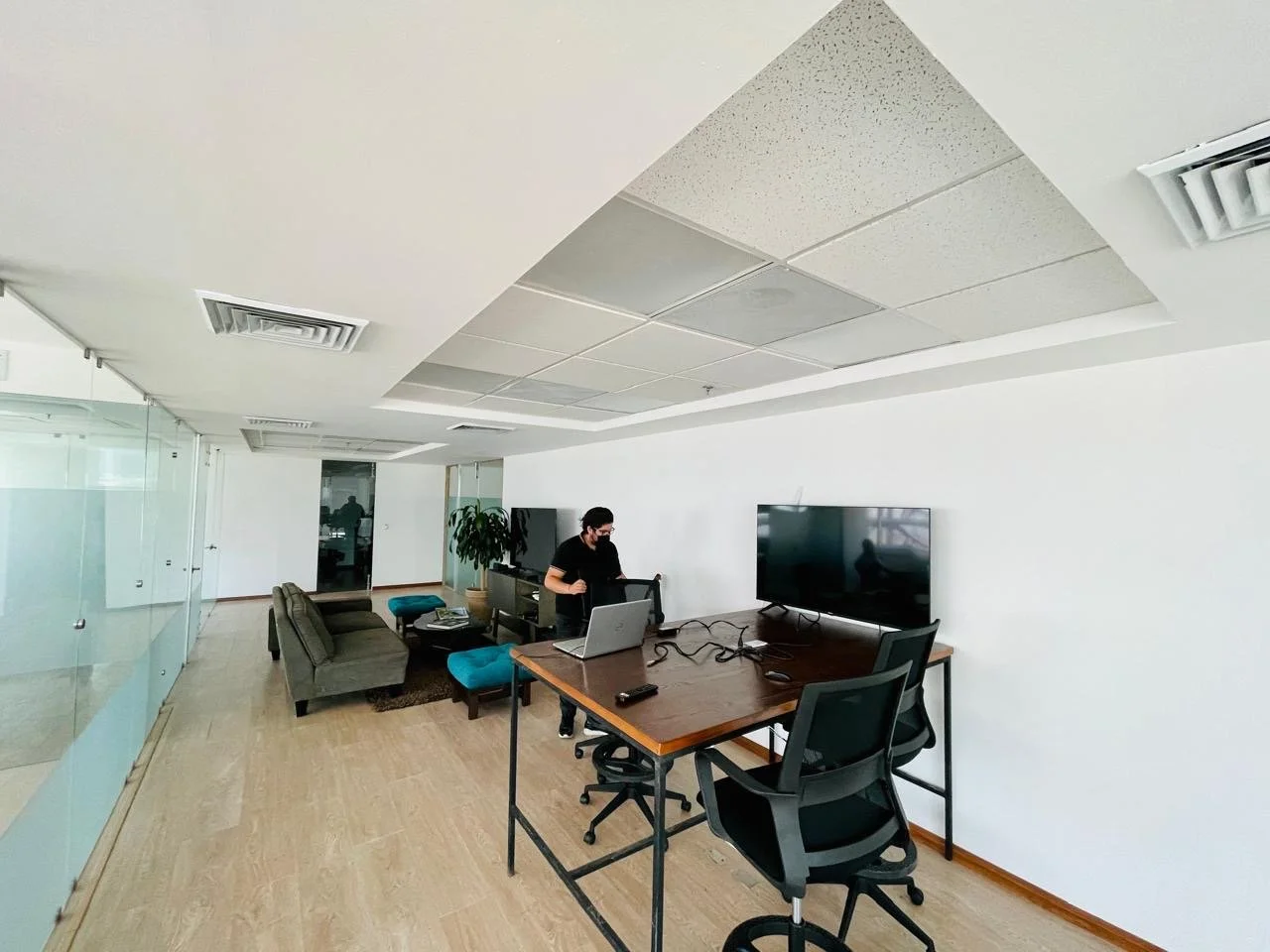 Office lounge with a person working at a table, TV screen, sofa, chairs, and potted plant.