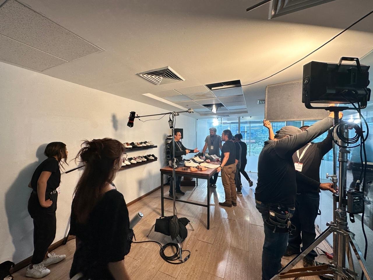 Behind-the-scenes view of a shoe product shoot in a modern room with a large window. Several crew members are setting up, adjusting equipment, and preparing shoes displayed on a table and shelves.