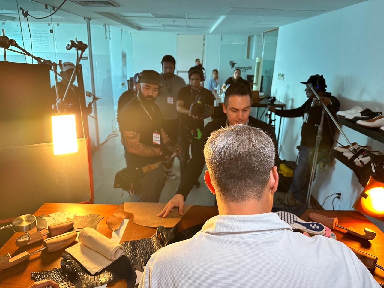 A team appears to be working on a leather craft project, with various leather pieces, tools, and fabric on a wooden table. Several people are observing or assisting, some wearing headphones and badges, in a well-lit craft studio.
