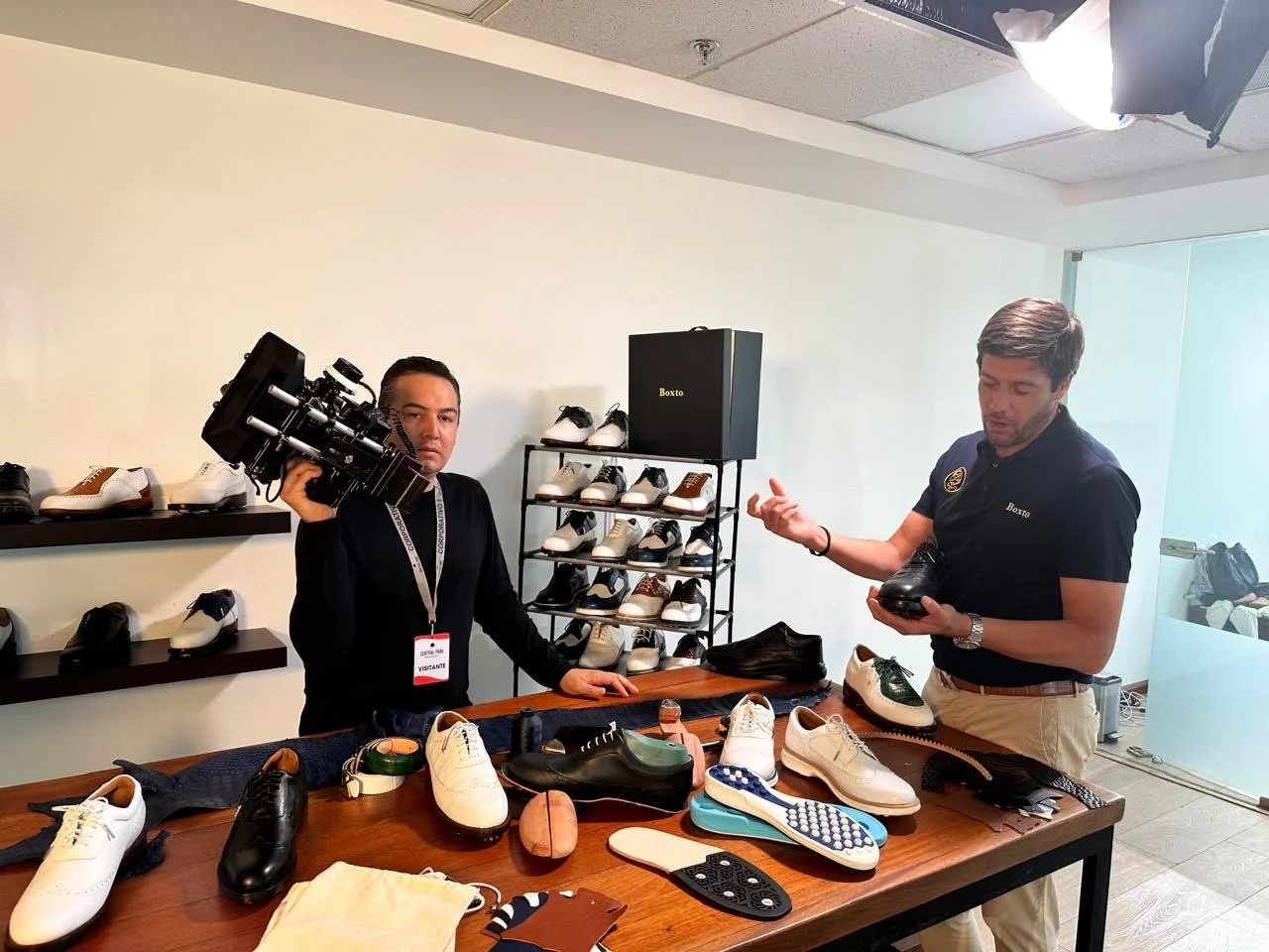 Two men in a shoe store or showroom, one holding a camera and the other inspecting a shoe. Merchandise includes various shoes and shoe care items on the table and shelves.