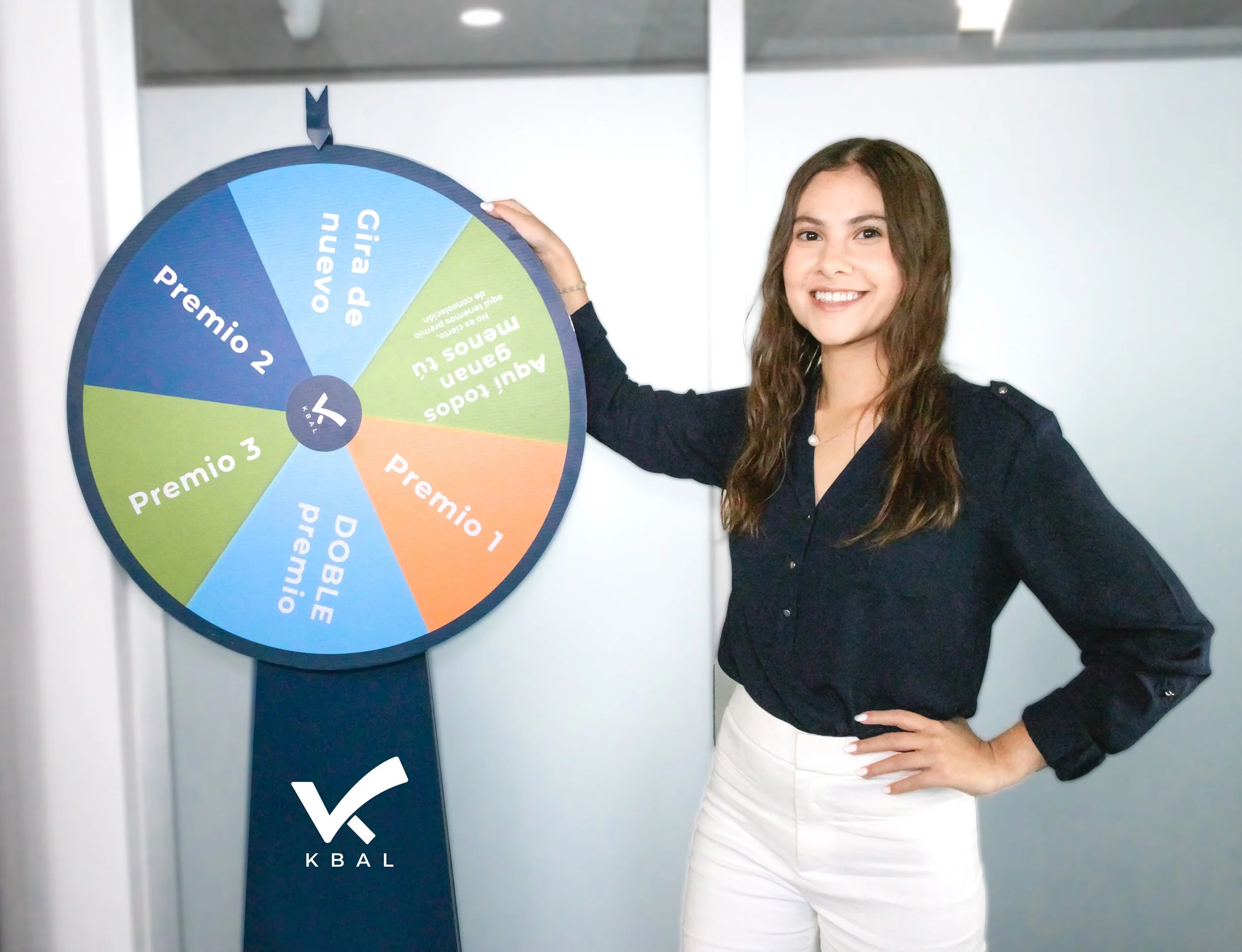 A woman with long brown hair, wearing a black blouse and white pants, smiling and posing with a colorful prize wheel labeled with different awards and graffiti, in an indoor setting.