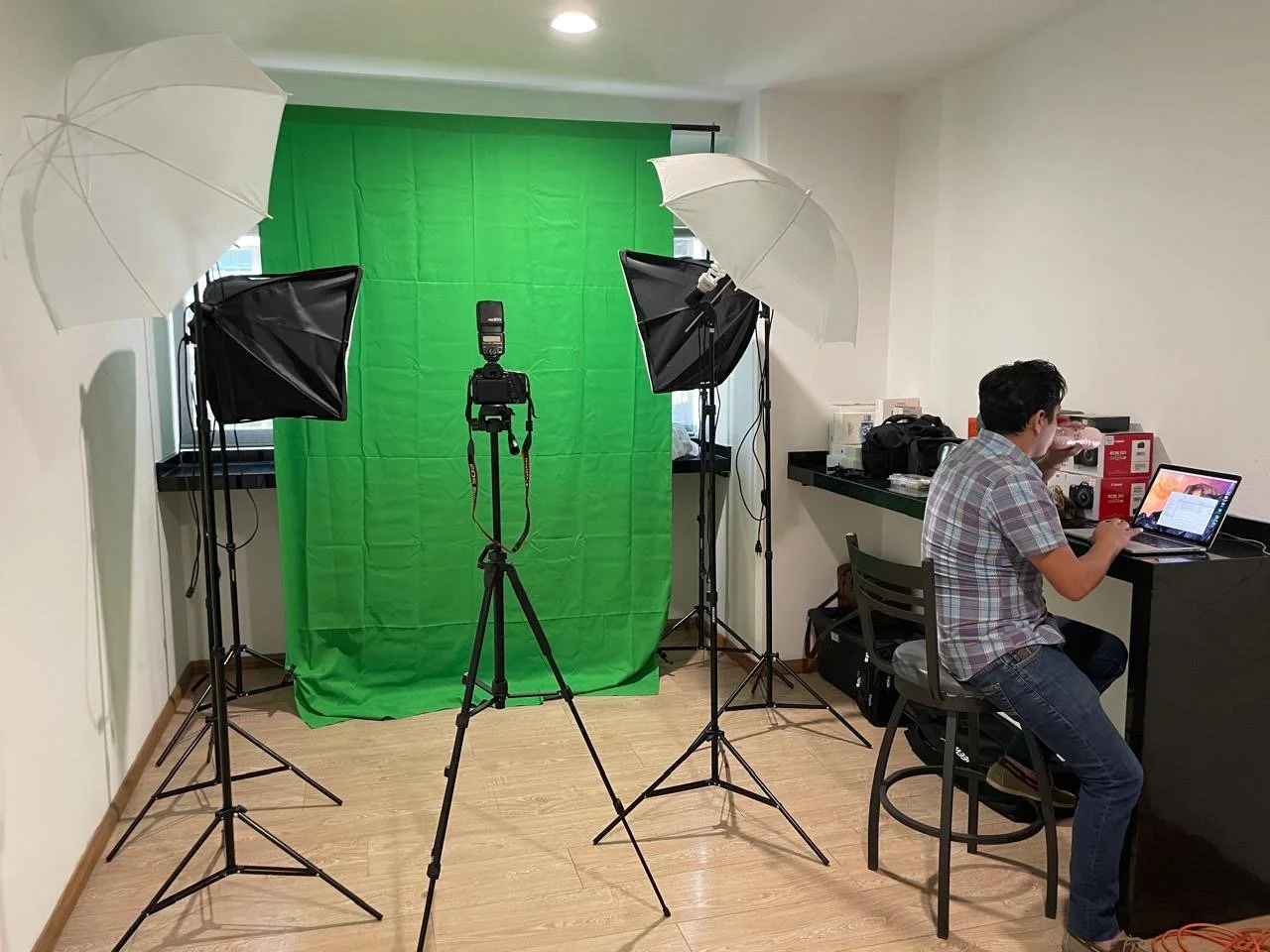 Home photography studio setup with green screen background, two softbox lights, two umbrella reflectors, and a person working at a desk with a laptop.