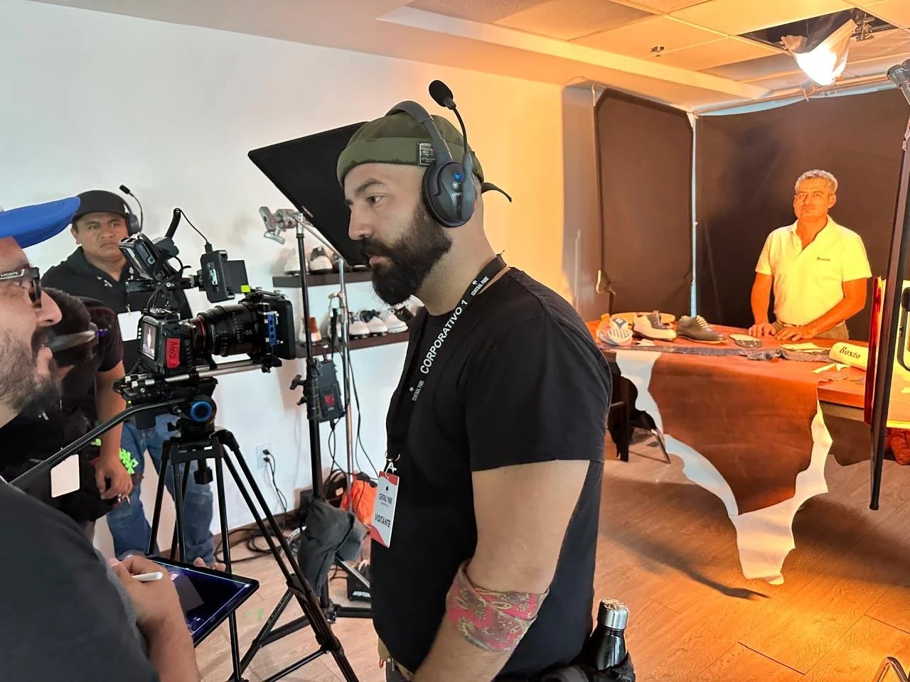 A man with a beard wearing a green beanie and headphones operates a professional video camera at a filming set. In the background, a man in a yellow shirt stands behind a table with shoes, with a black backdrop and lighting equipment.