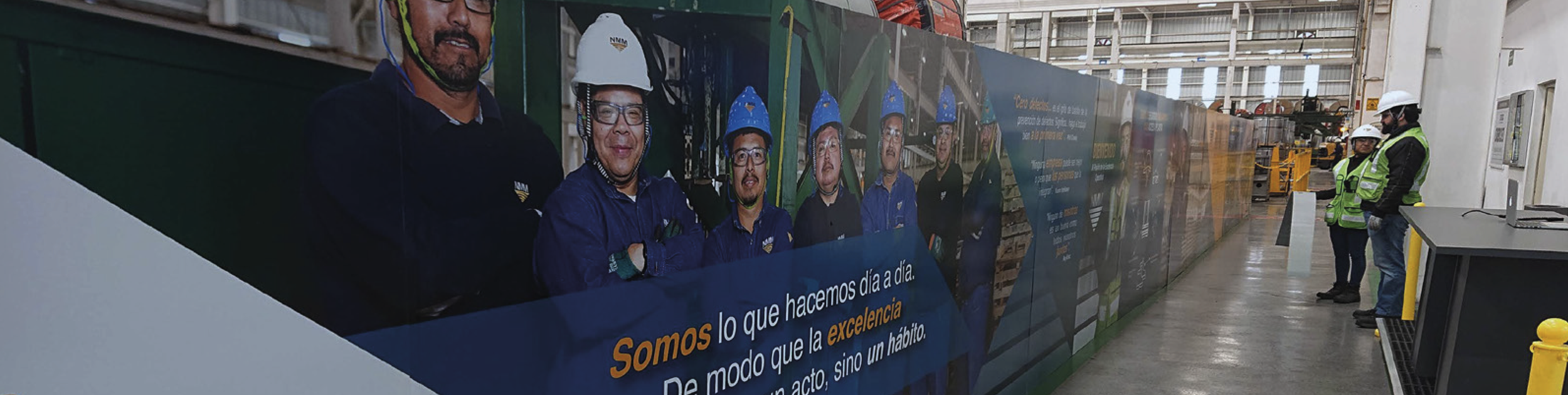 A group of workers wearing safety helmets and high-visibility vests observe a large poster inside an industrial or warehouse setting. The poster features several people in safety gear and some written text in Spanish.