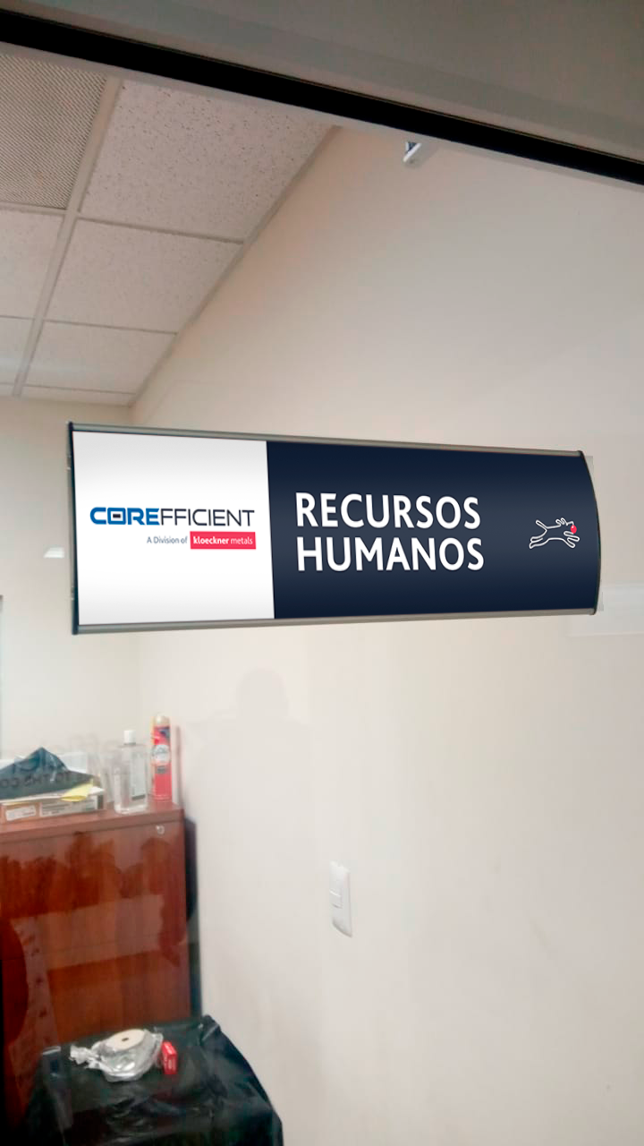 Indoor office hallway with a suspended sign that reads 'Recursos Humanos' and includes a company logo for CoreFicient, a division of Kloeckner Metals.