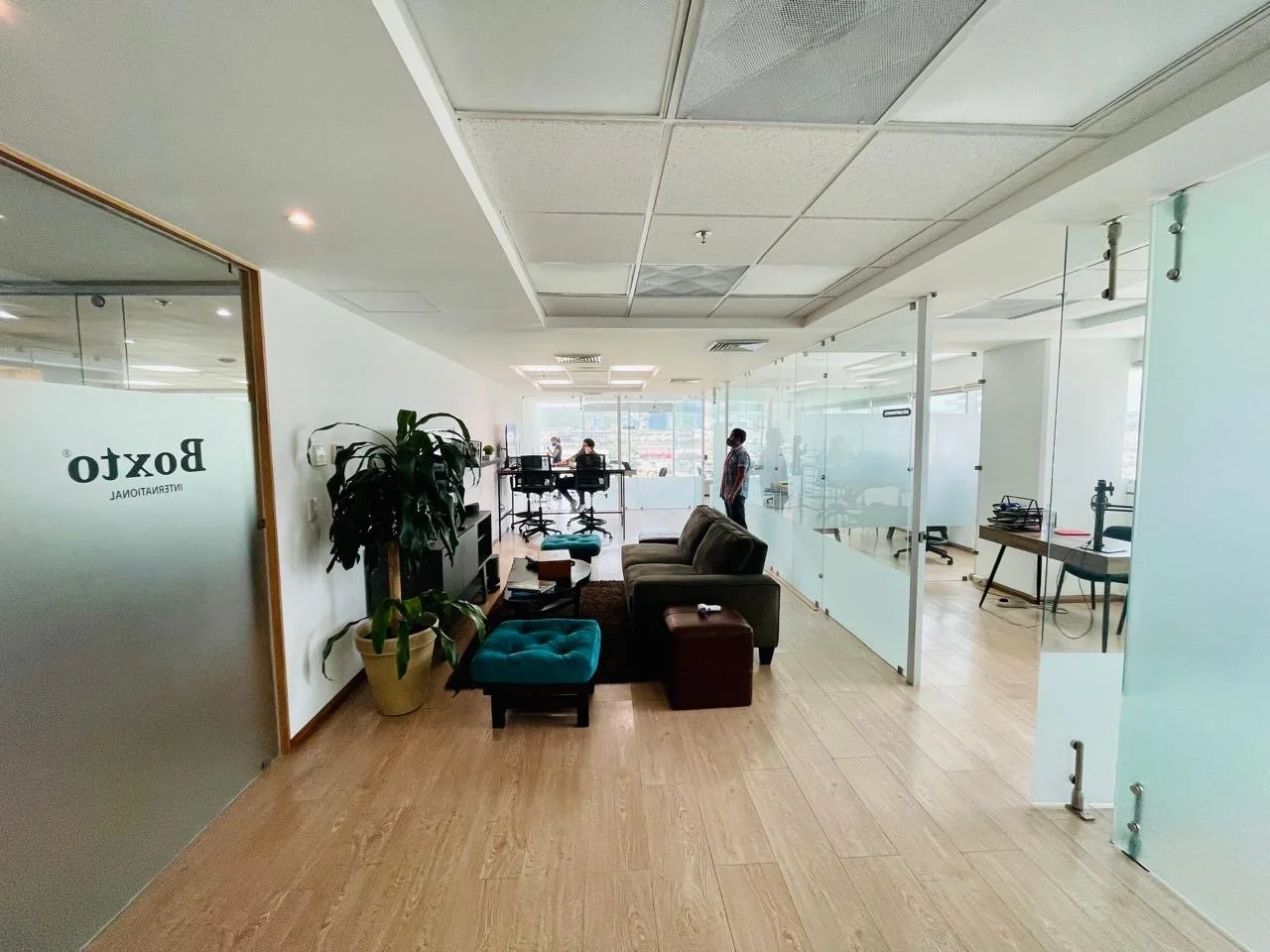 Office reception area with glass walls, seating area with black and gray sofas, potted plant, and people working at desks in the background.
