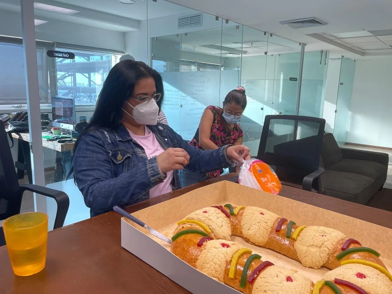 Two women wearing face masks, one with glasses and a denim jacket, and the other in a floral top, standing behind a table with a large cake decorated with colorful gummy candies, in an office setting.