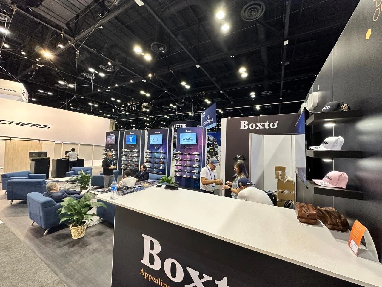 Trade show booth for Boxto featuring golf shoes, hats, and accessories with seating area and staff interacting with visitors.
