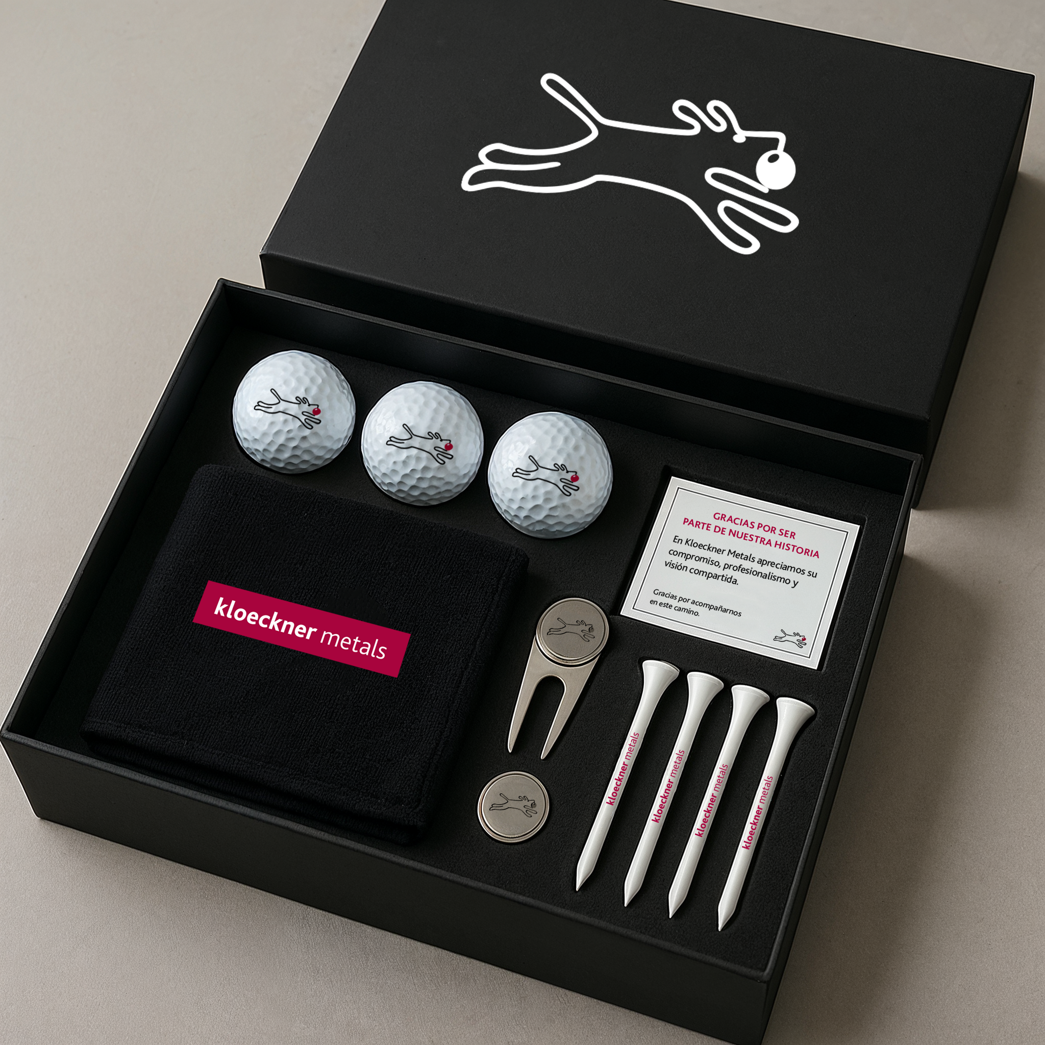 A black box containing a golf gift set with three golf balls, four golf tees, a small metal divot tool, a black cloth with 'kloeckner metals' written on it, and a thank you note in Spanish.
