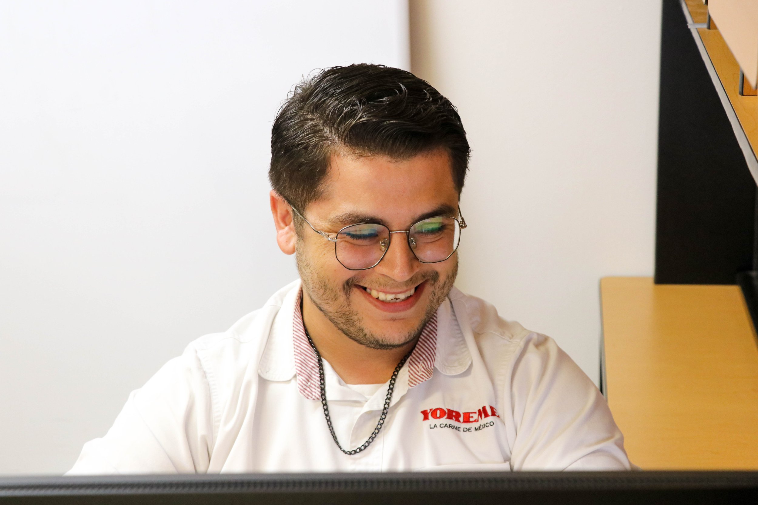 A smiling man with glasses looking at a computer screen, wearing a white shirt with red and black text and a chain necklace.