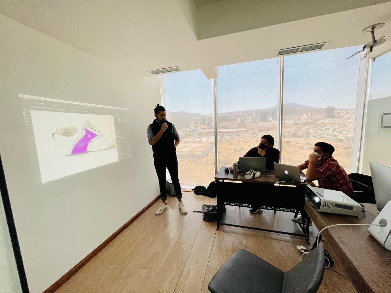 Person giving a presentation to two seated individuals in an office with a large window and a projector on the desk.