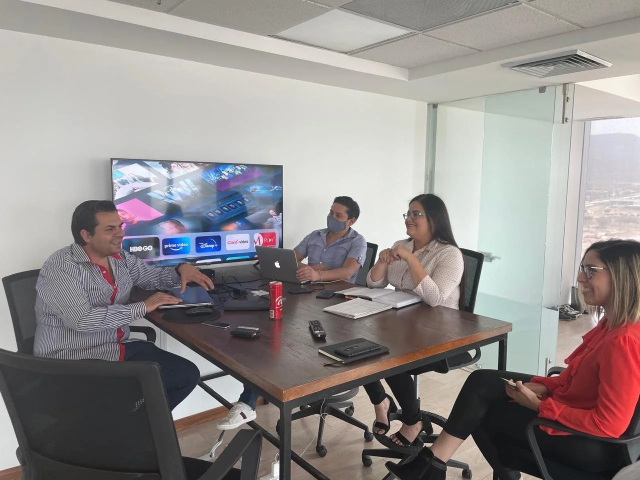 Four people in a conference room, three women and one man, sitting around a table with laptops, notebooks, and drinks, watching a presentation on a large screen.