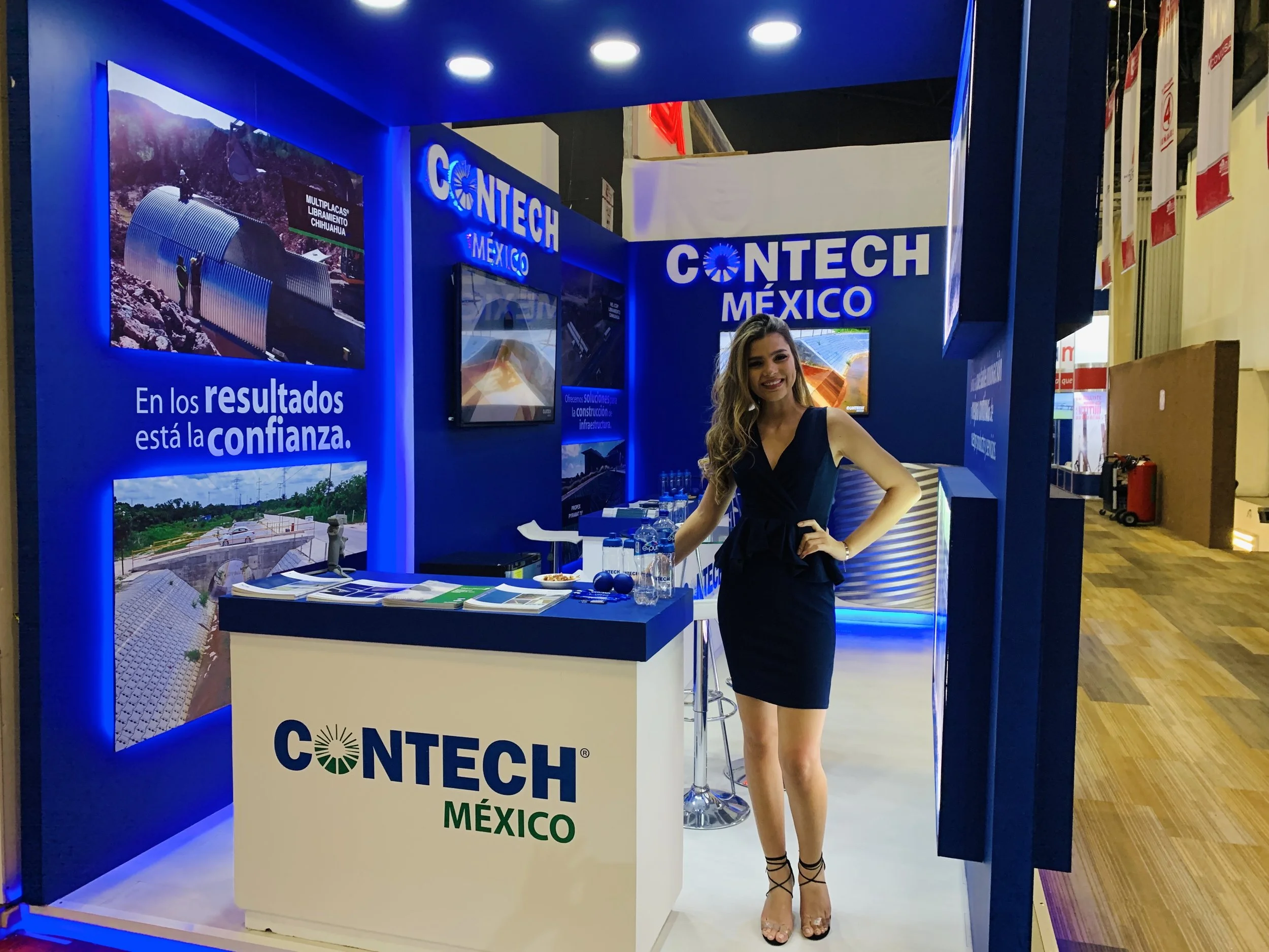A woman with long wavy hair in a black dress is standing at a trade show booth for CONTECH México. The booth has blue decor, posters, and a desk with brochures and water bottles. She is smiling and posing with one hand on her waist.
