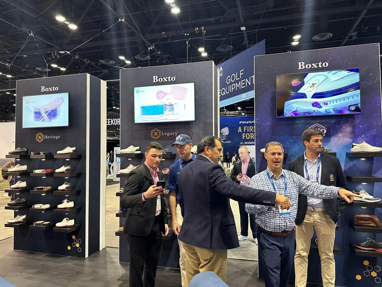 People at a trade show booth displaying golf shoes and equipment with the brand 'Boxto' and screens showing product images.