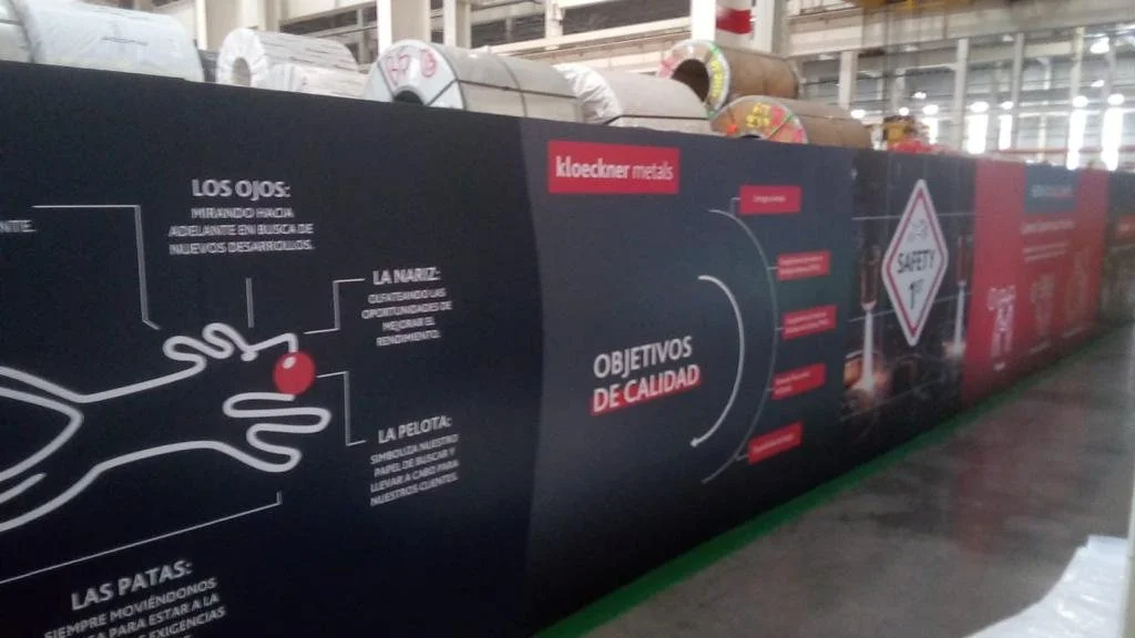 An industrial warehouse with storage racks holding large rolls of material. There are informational graphics and safety signs on the wall, and the environment appears spacious and brightly lit.