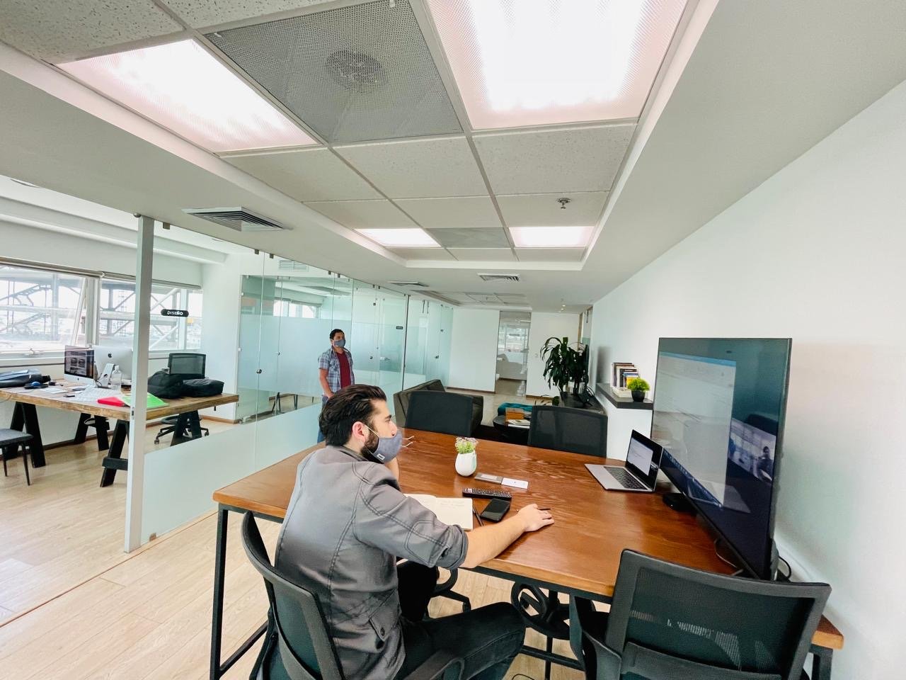 A man sitting at a desk in an office, wearing a mask and working on a computer, with two large screens and a laptop. In the background, another person wearing a mask stands near a glass partition. The office has bright lighting, white walls, and wood