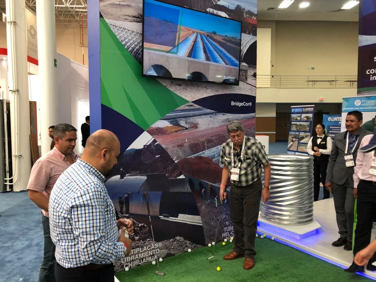 People at a trade show exhibit with a large display of pipes, a screen showing construction images, and a golf putting green with golf balls and cups.