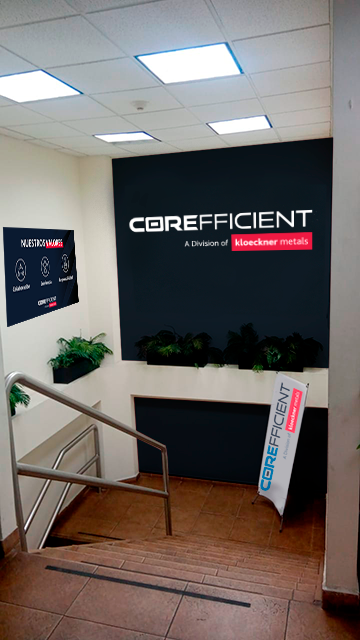 Indoor staircase leading down to a company reception area with signage for 'Correctence, a division of Kloeckner Metals,' featuring potted plants and informational posters on the wall.