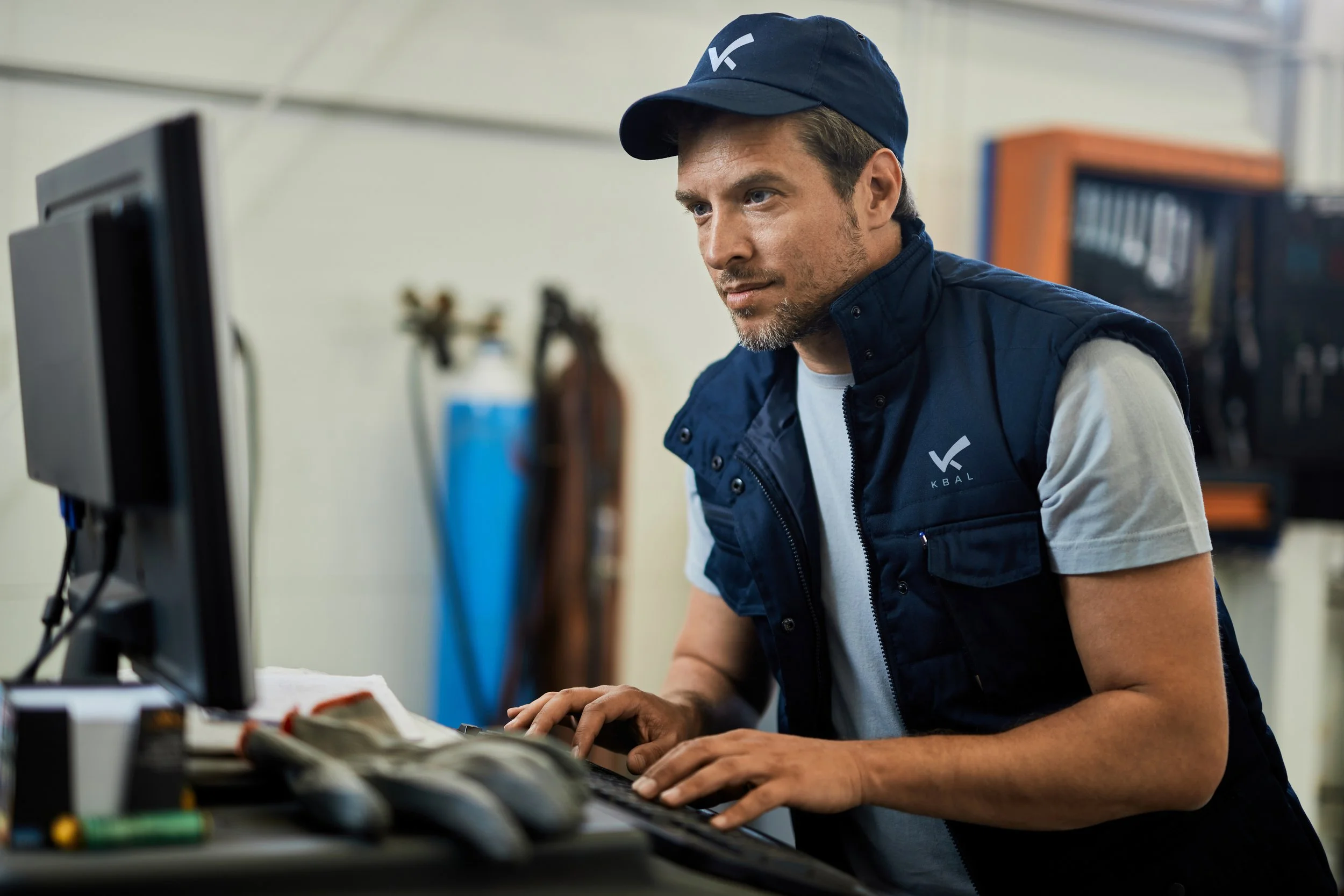 A man wearing a blue cap and vest with the KBAL logo working on a computer in an industrial setting.
