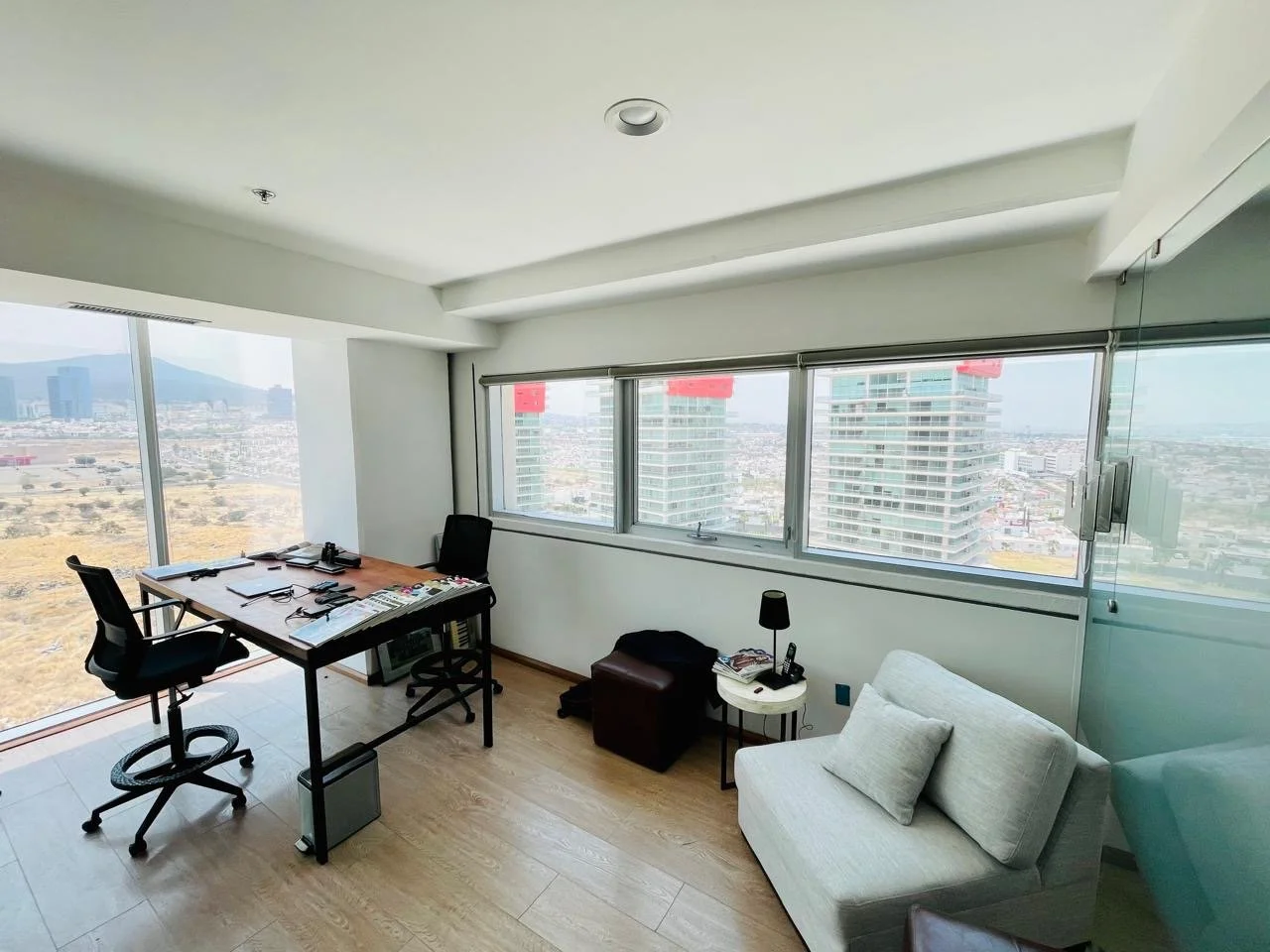 Office room with a desk, two chairs, a sofa, and a large window showing cityscape and tall buildings.