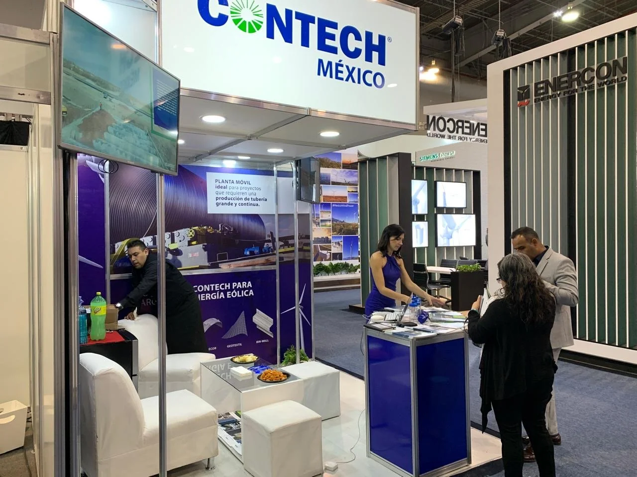 Trade show booth for CONTECH MEXICO with displays about wind energy technology, a seating area with white chairs, and three people engaging in conversation and work at a counter.