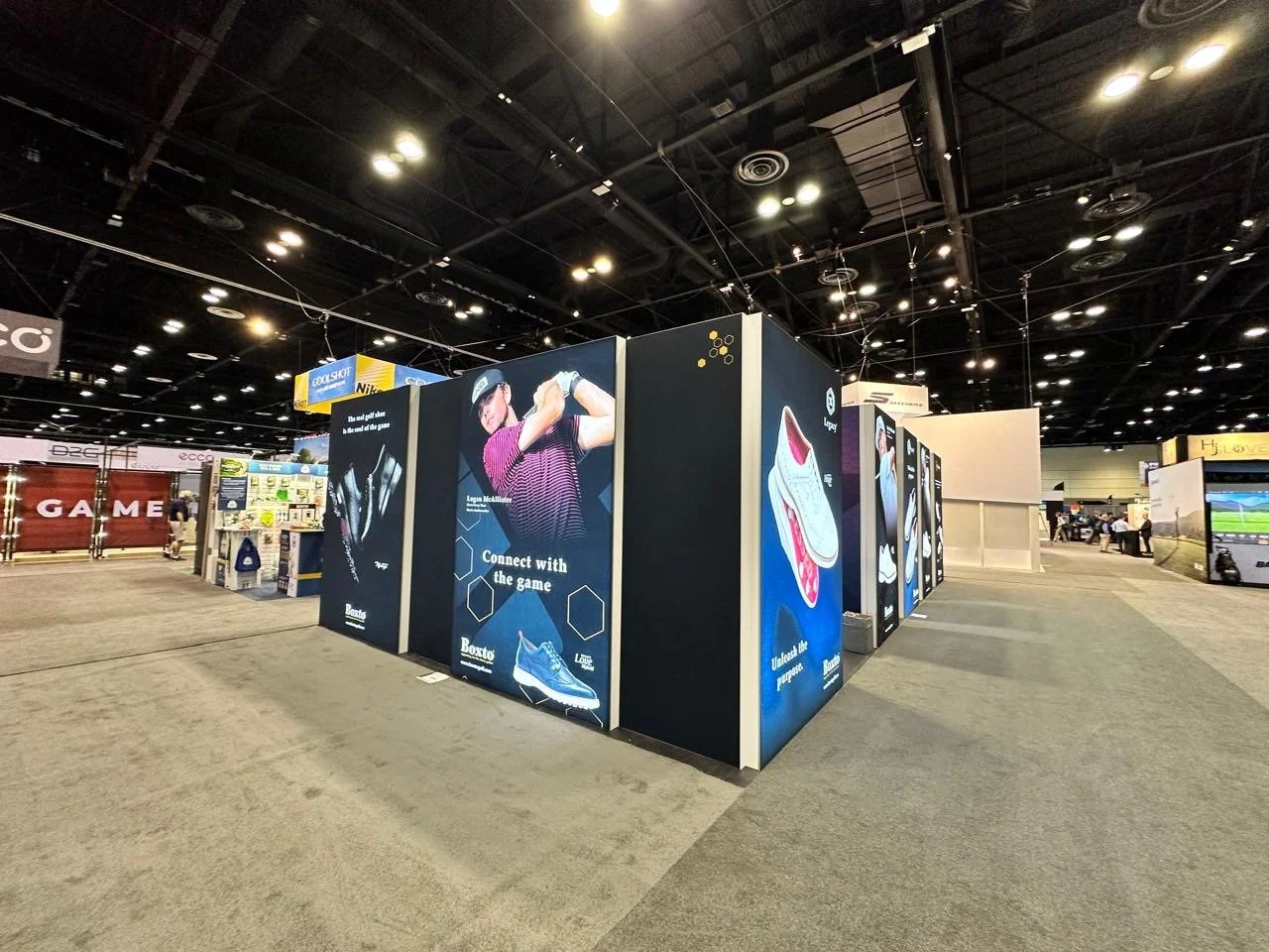 Indoor trade show with various advertising banners for sports and fashion brands, including a large banner featuring a golfer and athletic shoes, and a sports field in the background.