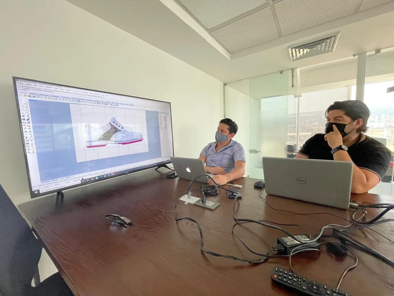 Two men sit at a conference table, both using laptops, watching a large display screen showing a 3D design of a shoe. Both men wear masks in a bright office with large windows.