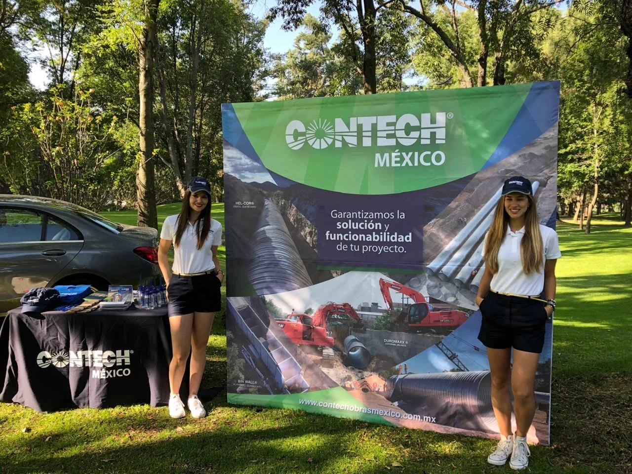 Two women standing in front of a CONTECH Mexico promotional banner at an outdoor event. One woman is standing next to a table with brochures and water bottles, and both women are smiling and wearing white polo shirts, black shorts, and caps.